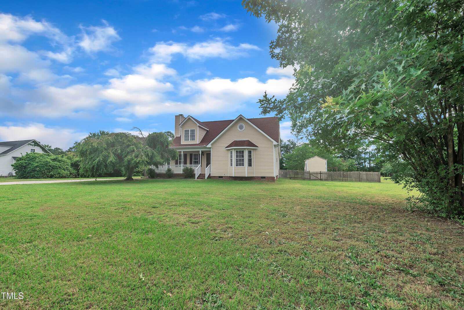 A beige two-story house with a red roof sits on a large, grassy yard with trees and a driveway under a partly cloudy sky. A fenced area and another building are visible in the background.