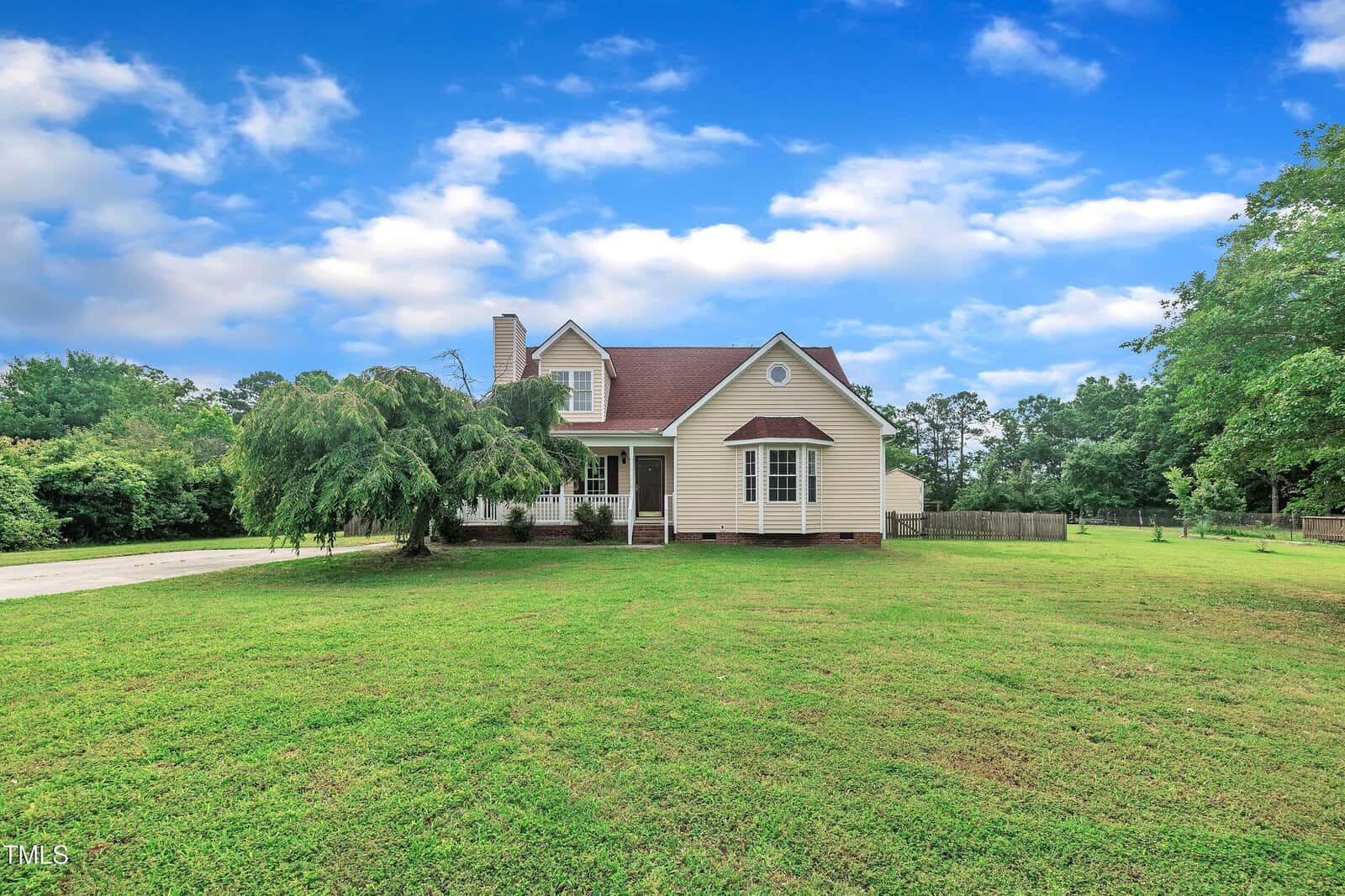 A charming yellow house with a red roof sits on a large, green lawn under a partly cloudy blue sky, surrounded by trees and a driveway on the left side.