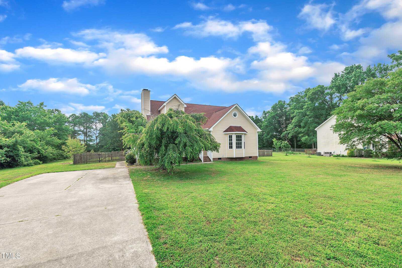 A single-story house with beige siding and a red roof sits beside a large concrete driveway, surrounded by a green lawn and trees under a blue sky with scattered clouds.