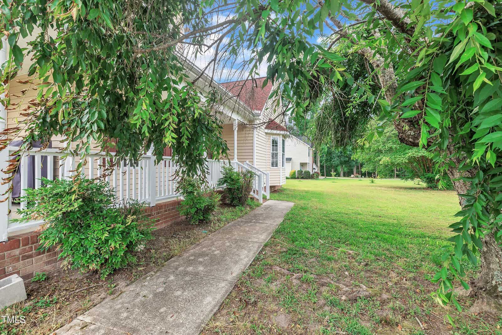 A concrete walkway leads to a house with a brick foundation and white porch railing, surrounded by green bushes and trees, opening onto a grassy yard under a blue sky.