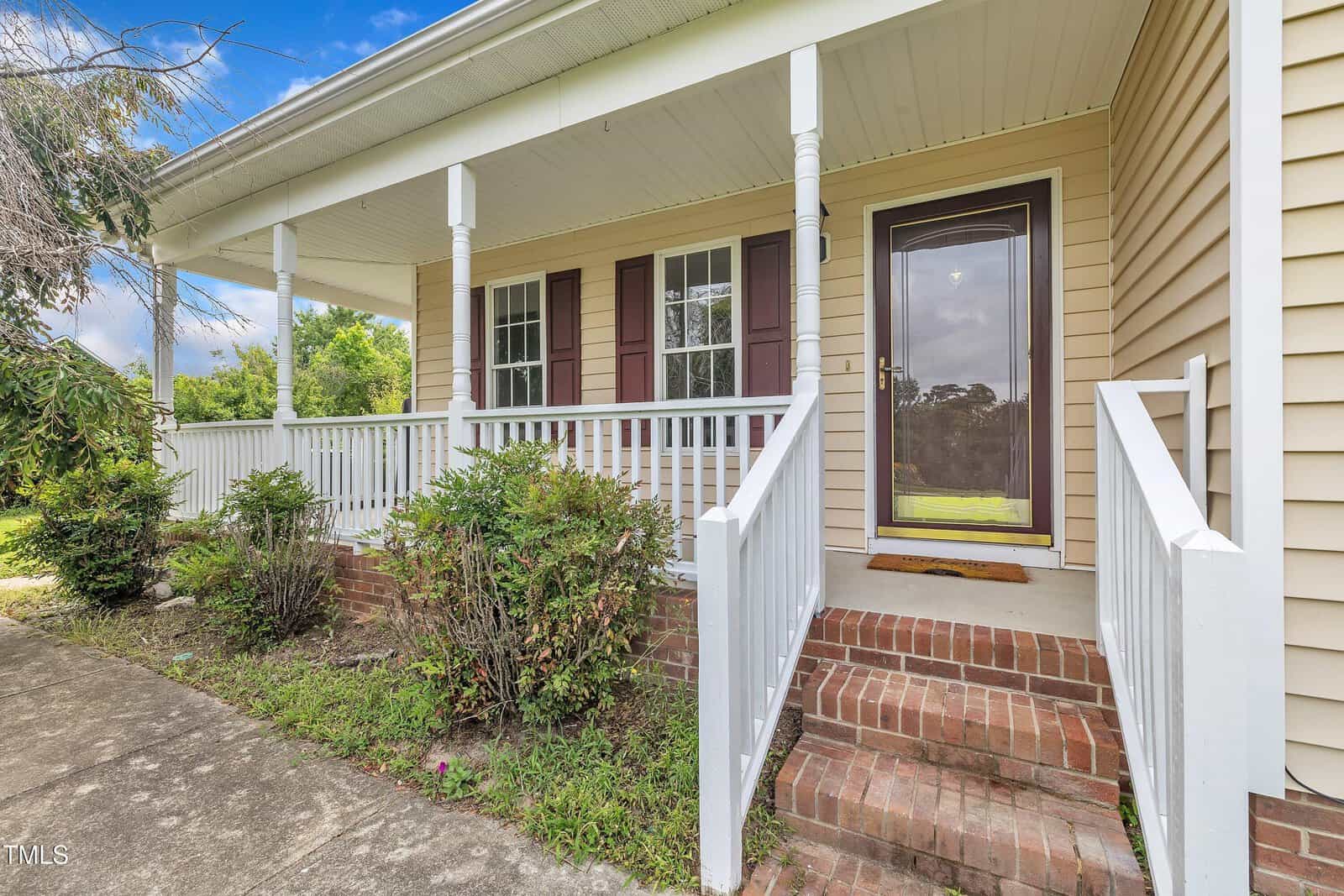 A beige house features a covered front porch with white railings, brick steps, a glass storm door, and two windows with brown shutters. Shrubs line the walkway leading to the entrance.