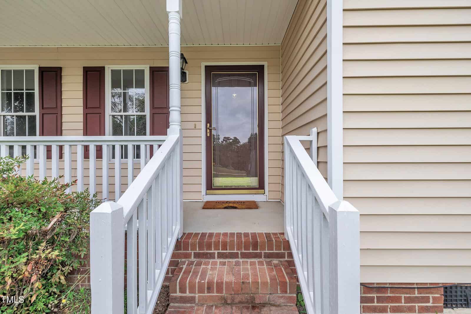 A front porch with beige siding, white railings, brick steps, and a glass storm door. Two windows with brown shutters are on the left, and green foliage is near the steps.