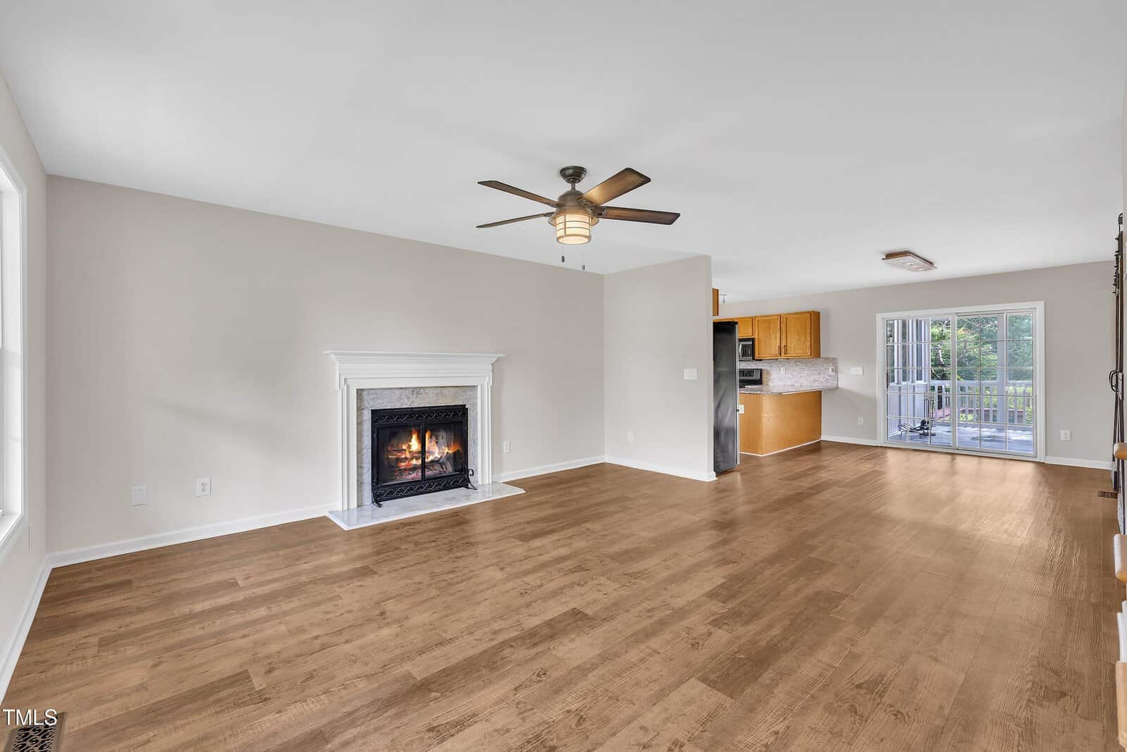 Spacious living area with hardwood floors, ceiling fan, lit fireplace, and neutral walls. Open layout connects to a kitchen and dining area with large windows and a door to a deck outside.