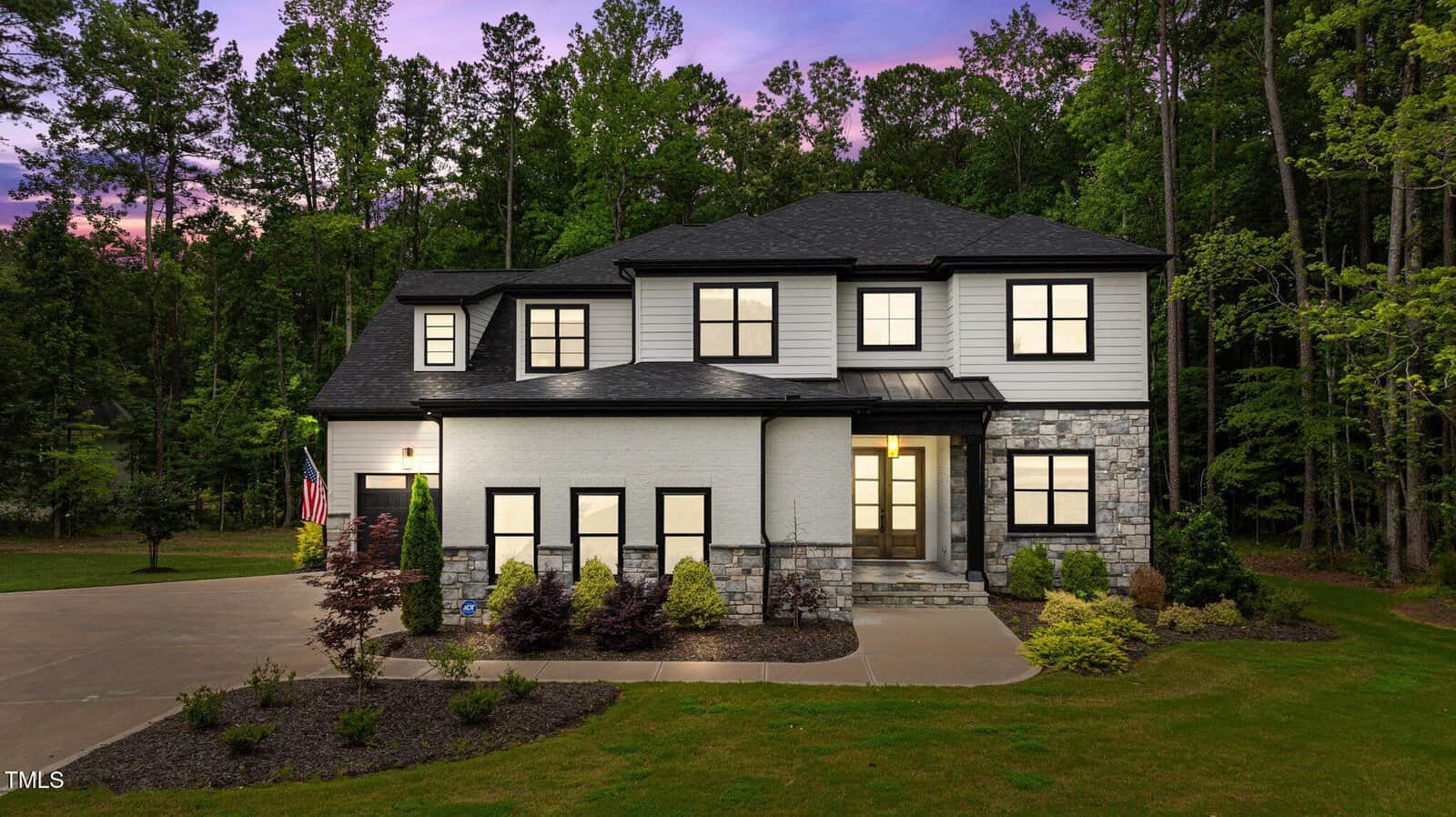Modern two-story house with white and stone exterior, large windows lit from inside, black roof, and landscaped yard; surrounded by trees at dusk with a purple sky in the background.