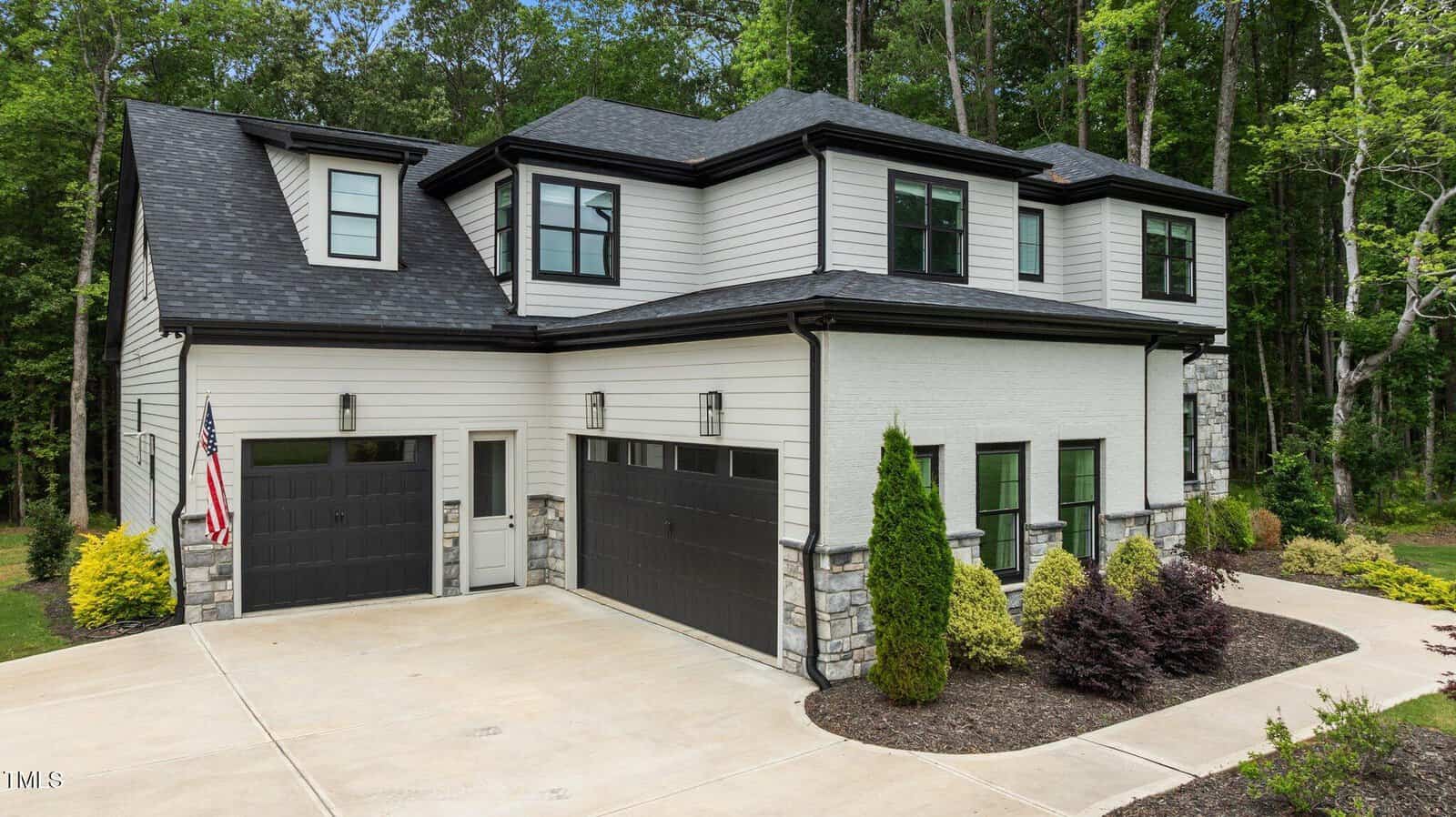 Modern two-story house with white siding, black trim, and black garage doors, set among trees. The driveway is concrete, and the front yard features landscaped bushes and an American flag by the entrance.