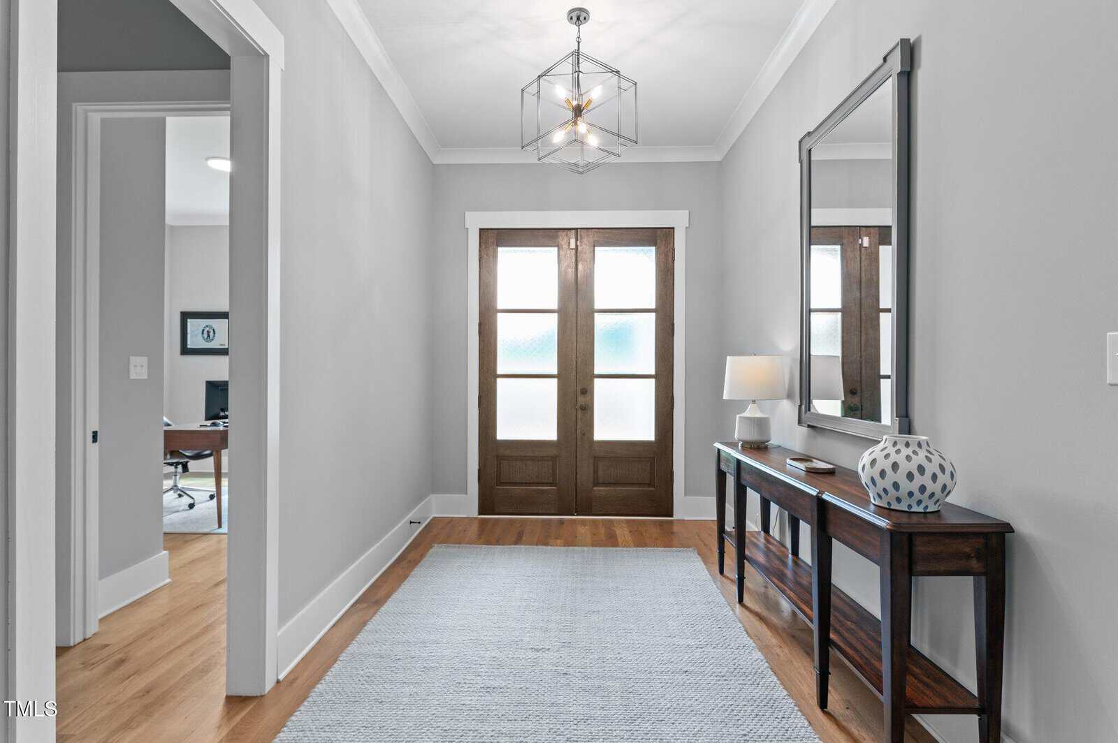 A bright, modern entryway with double wooden front doors, a large rectangular mirror above a dark wood console table, a table lamp, and a decorative vase. Light gray walls and hardwood floors create an inviting atmosphere.
