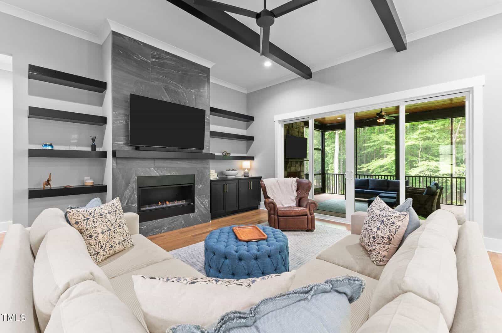 A modern living room with a sectional sofa, blue ottoman, fireplace with TV above, open shelves, and large windows showing a covered porch and trees outside. A leather chair and side table sit near the window.