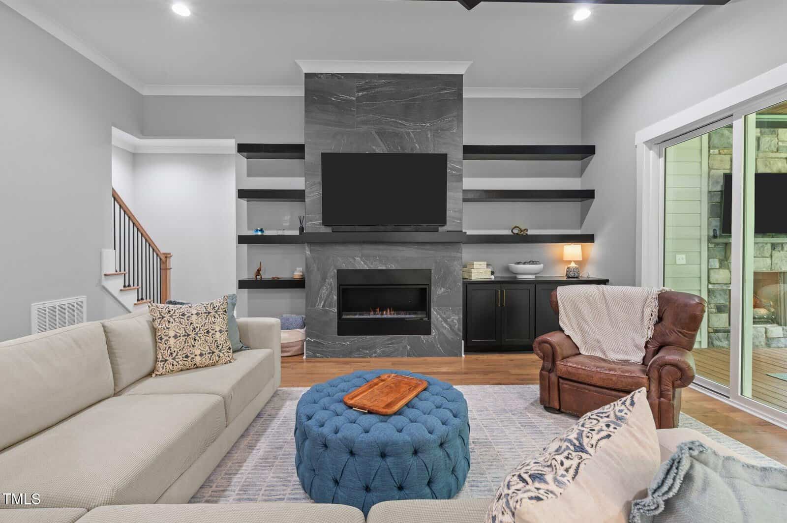 Modern living room with light sofas, a brown leather armchair, blue tufted ottoman, built-in shelves, a large TV above a sleek fireplace, and sliding glass doors leading outside. Decor is neutral with cozy accents.