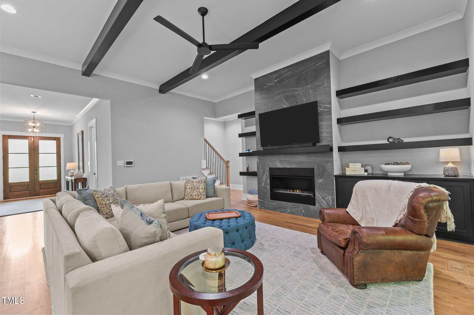 Modern living room with a beige sectional sofa, leather armchair, blue ottoman, and built-in shelving around a fireplace and wall-mounted TV. Large ceiling fan and wood beam accents are visible. Natural light enters from the entryway.