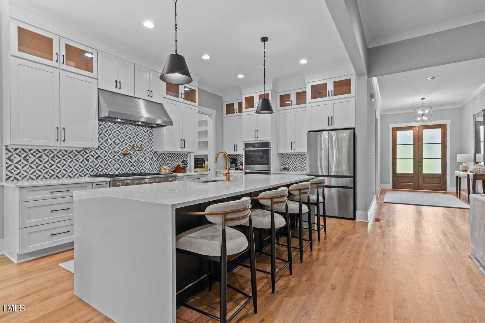 Modern kitchen with white cabinets, stainless steel appliances, patterned tile backsplash, large island with sink, and four barstools. Pendant lights hang above the island, and wood flooring extends throughout the space.
