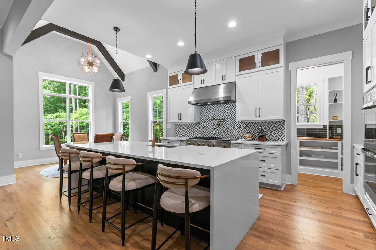 Modern kitchen with a large island, four barstools, pendant lights, white cabinets, patterned tile backsplash, stainless steel appliances, wood floors, and large windows. An open dining area and pantry are visible in the background.