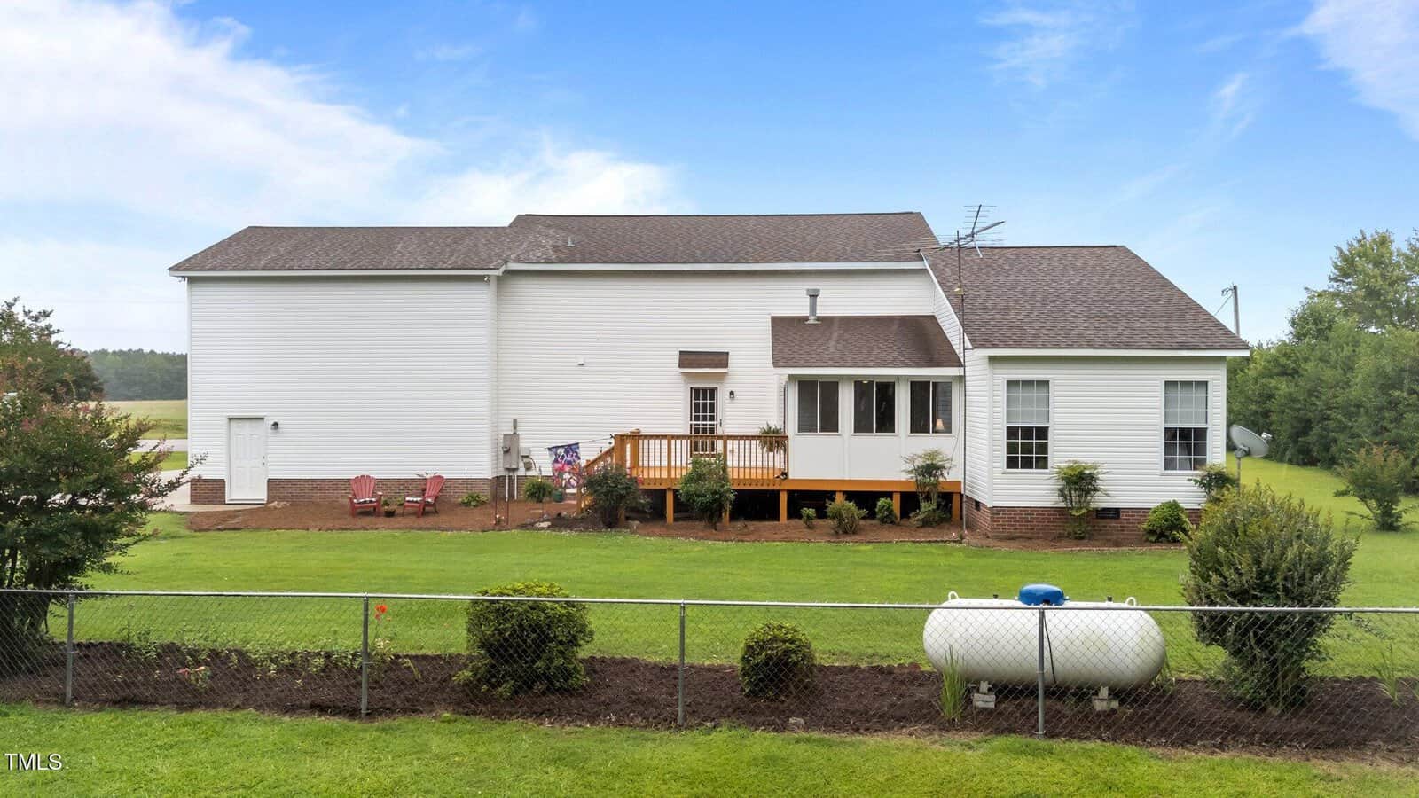 A white single-story house with a screened porch, a wooden deck, and a brown shingle roof, sits in a grassy yard with shrubs and a propane tank, enclosed by a chain-link fence under a blue sky.