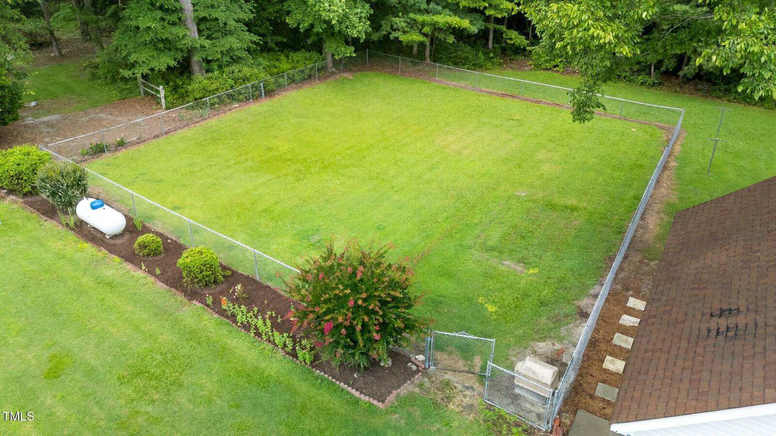 A fenced backyard with green grass, a small garden bed, and a white propane tank on the left. The area is bordered by trees and a house roof is visible in the bottom right corner.
