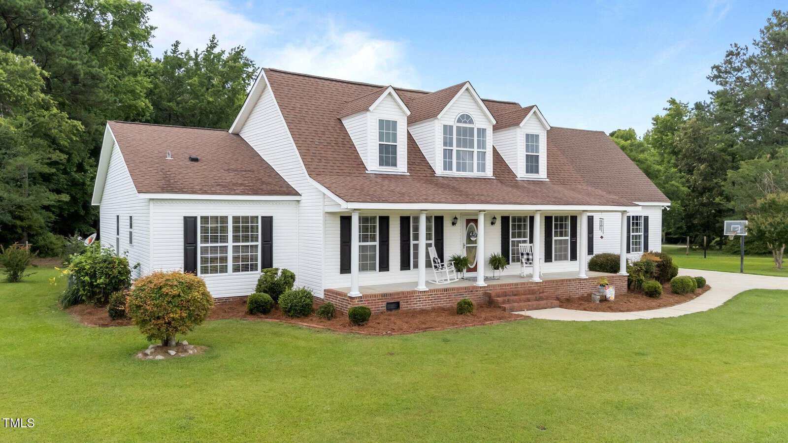 A white two-story house with brown roof shingles, dormer windows, black shutters, a covered front porch, and neatly trimmed bushes, surrounded by a large, well-maintained green lawn under a blue sky.