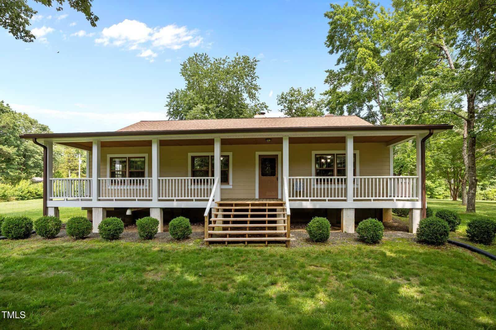 A charming one-story house at 3044 Rock Springs Church Road in Creedmoor features a large covered front porch with white railings, beige siding, a brown roof, and wooden steps. Lush green lawn, shrubs, and trees complete the inviting setting.