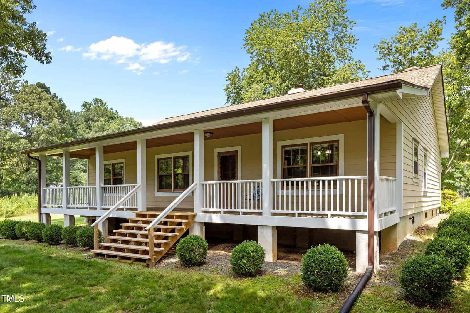A single-story beige house with a large covered front porch, white railings, wooden steps, and neatly trimmed bushes along the front, surrounded by green grass and trees under a blue sky.