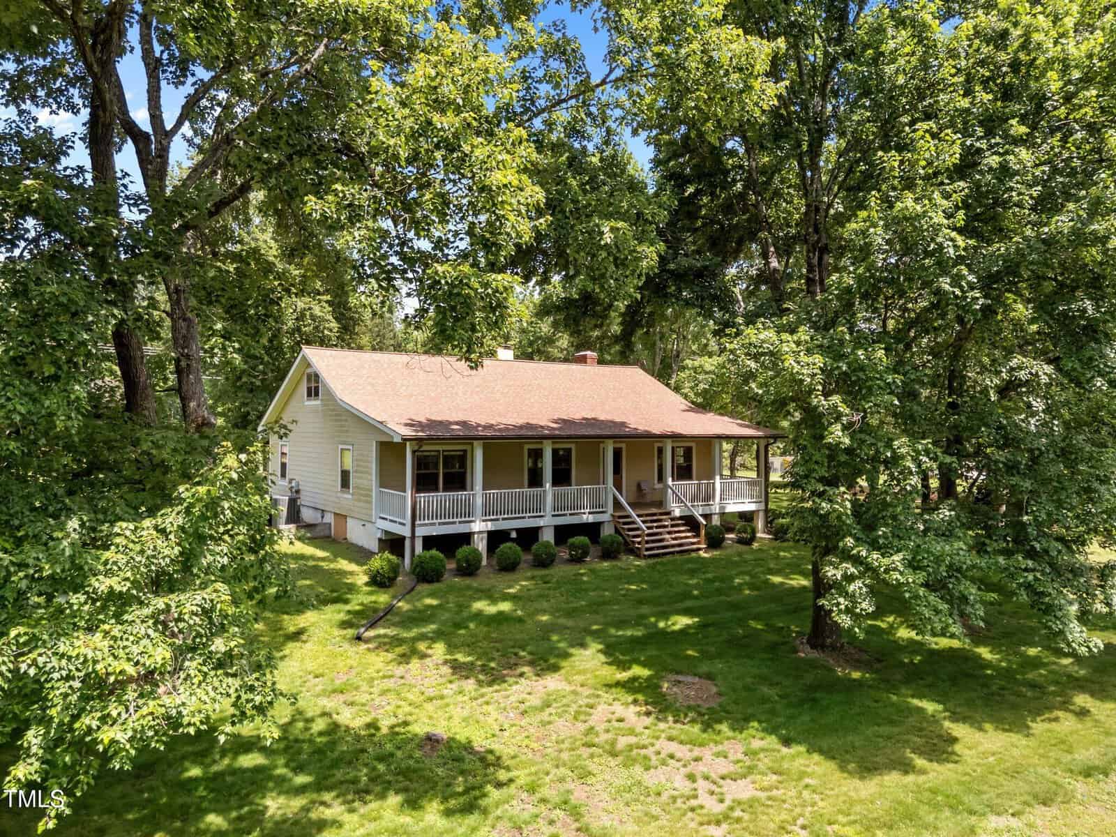 A single-story house with a large covered front porch and stairs, situated on a grassy lawn and surrounded by tall trees under a clear blue sky.