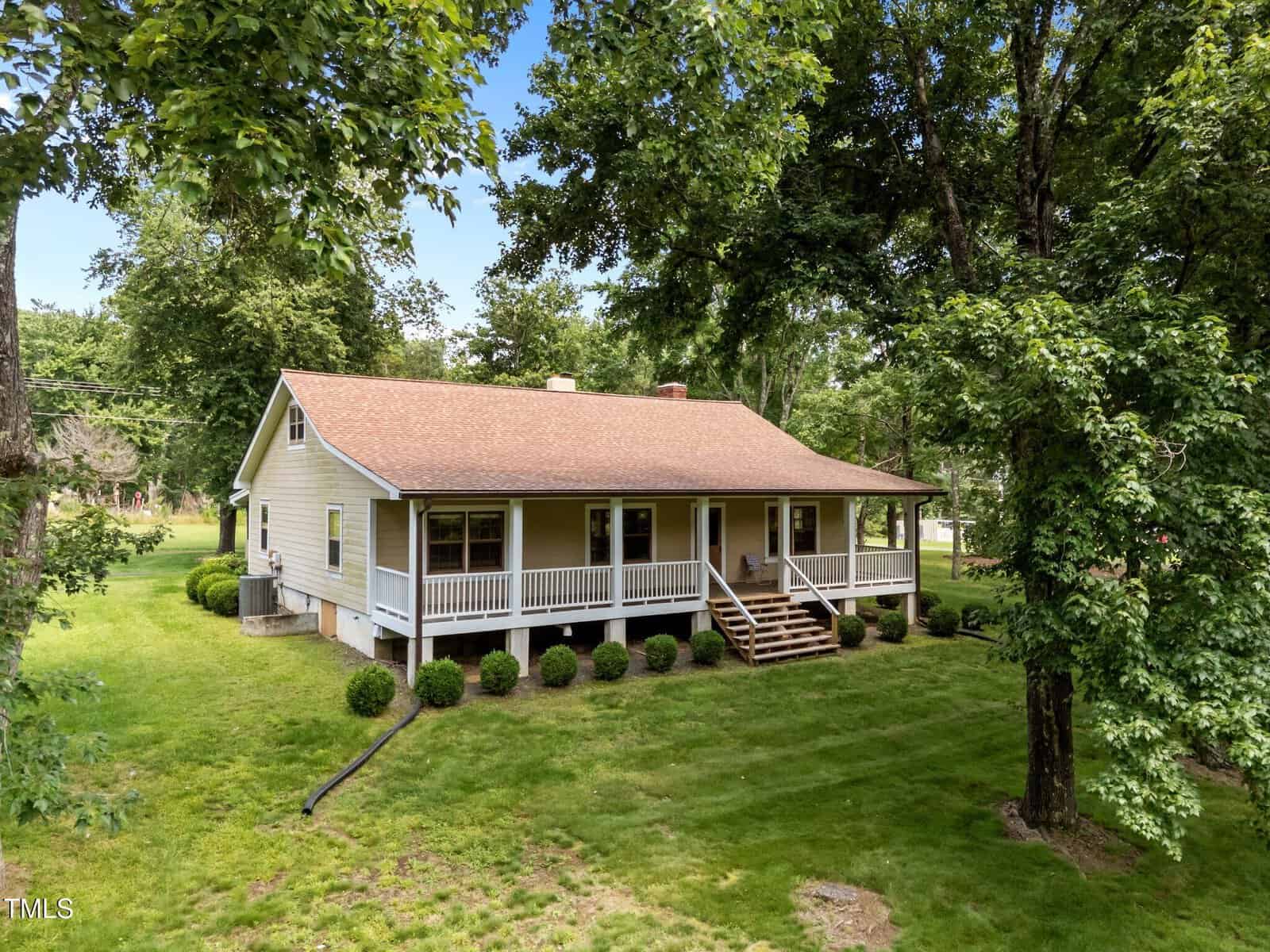 A single-story house with a covered front porch, tan siding, and a brown roof sits amid a well-kept lawn and mature trees, with stairs leading up to the porch and bushes along the foundation.