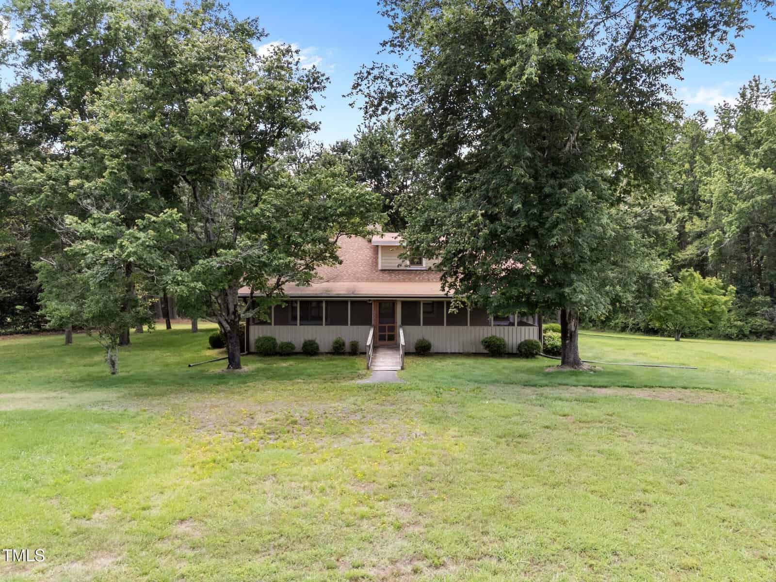 A tan, two-story house with a covered front porch sits behind two large trees in a spacious grassy yard surrounded by woods. A walkway leads from the porch to the foreground.
