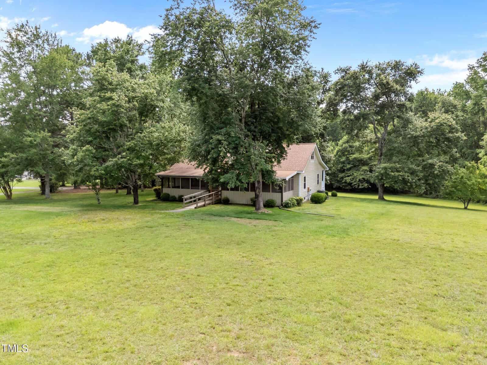 A single-story house with a covered front porch and ramp sits surrounded by a large grassy yard and mature trees under a blue sky with scattered clouds.