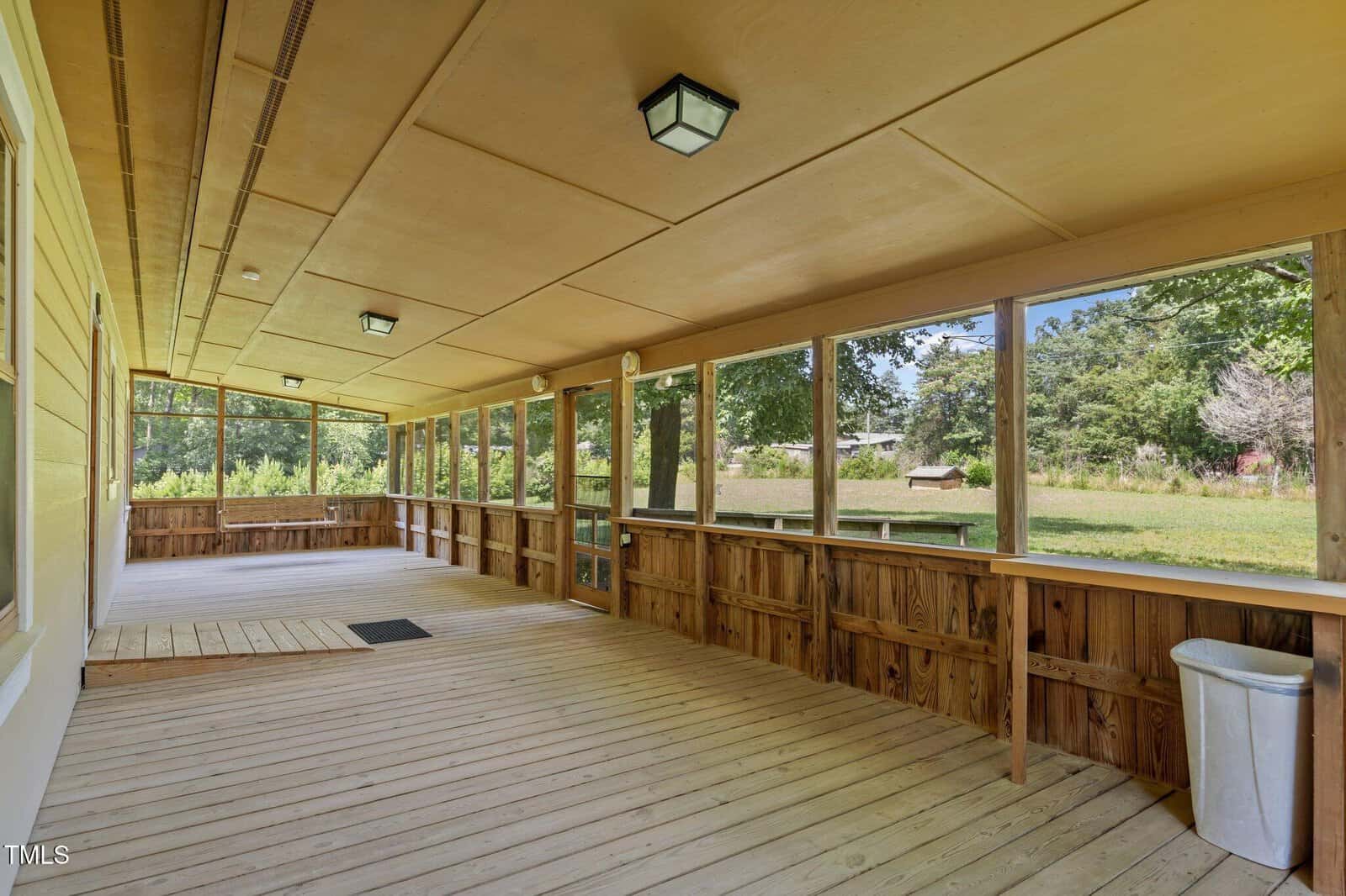Screened-in wooden porch with a light wood floor and ceiling, lights overhead, and partial open views of a sunny grassy yard with trees. A trash can sits in the right corner near the screen.