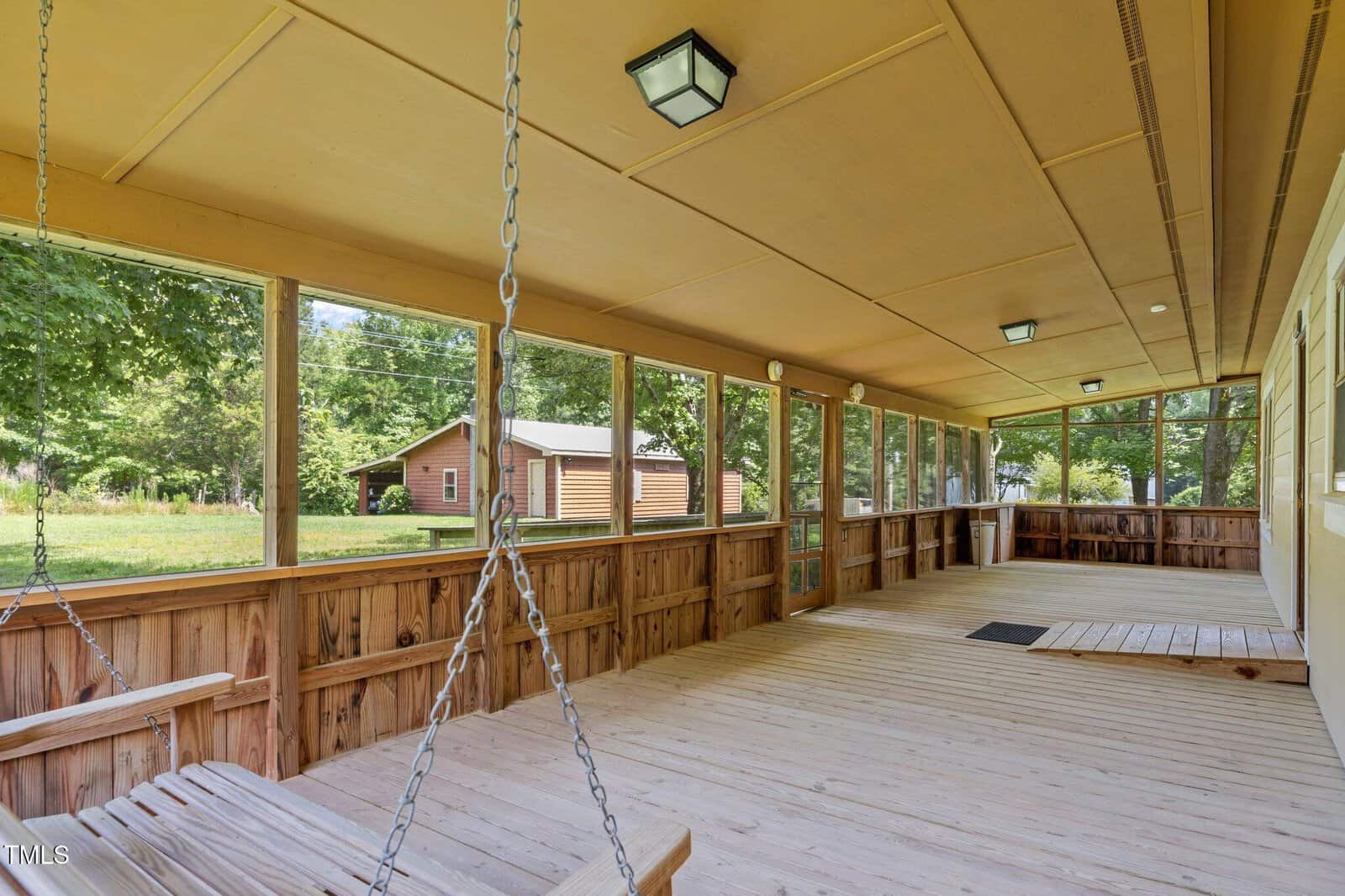 Spacious screened-in porch with wooden flooring, ceiling lights, and two porch swings. The view shows trees and a wooden cabin outside.
