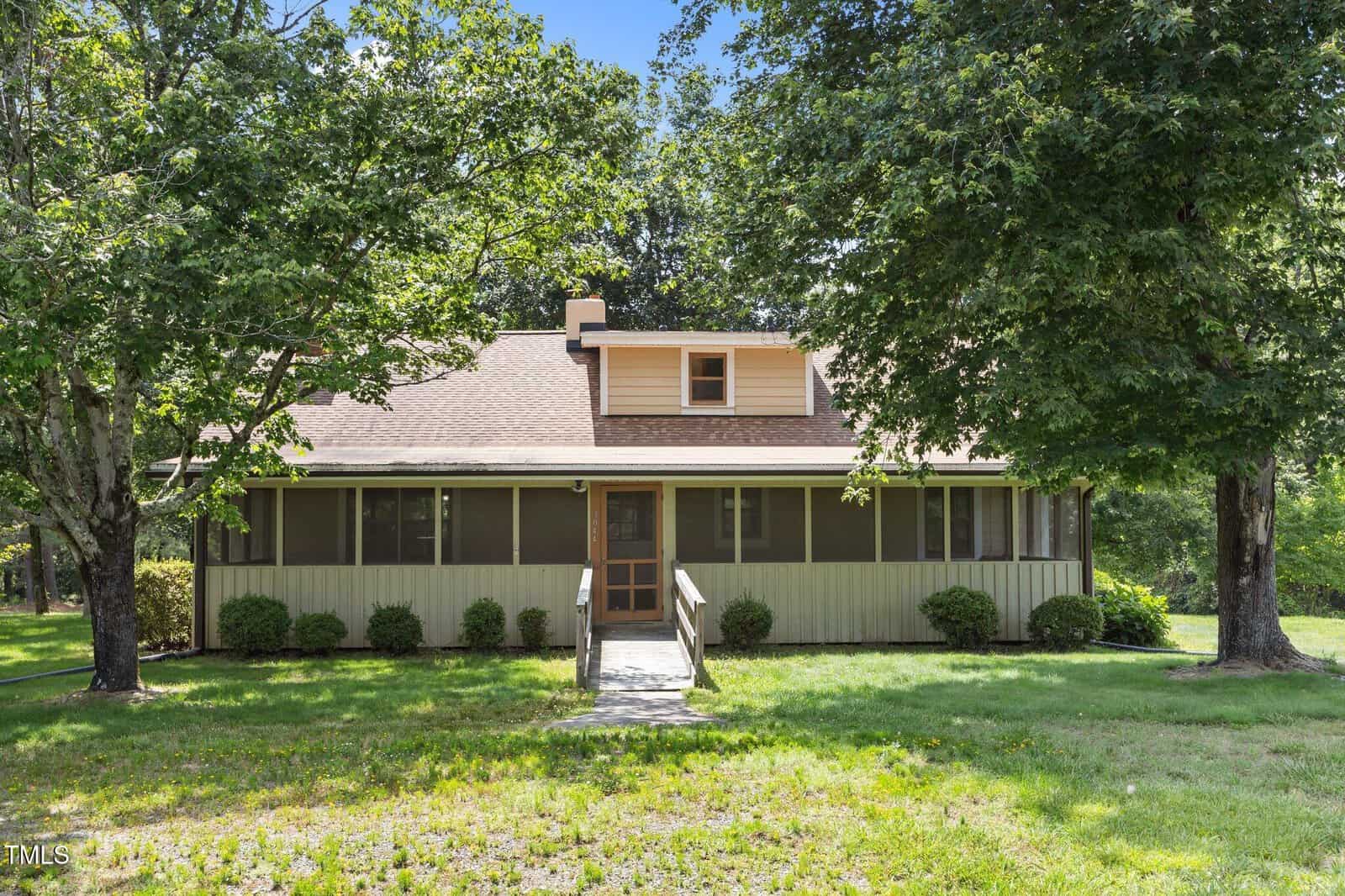 A one-story house with a screened front porch, wooden ramp, dormer window, and tan siding, surrounded by green grass, bushes, and large leafy trees under a clear blue sky.