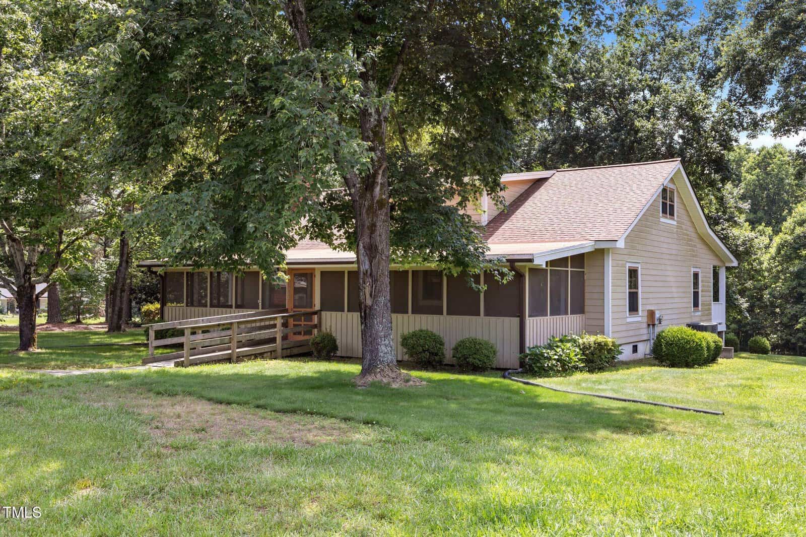 A tan, single-story house with a screened-in porch and ramp sits on a grassy lawn surrounded by trees and shrubs on a sunny day.