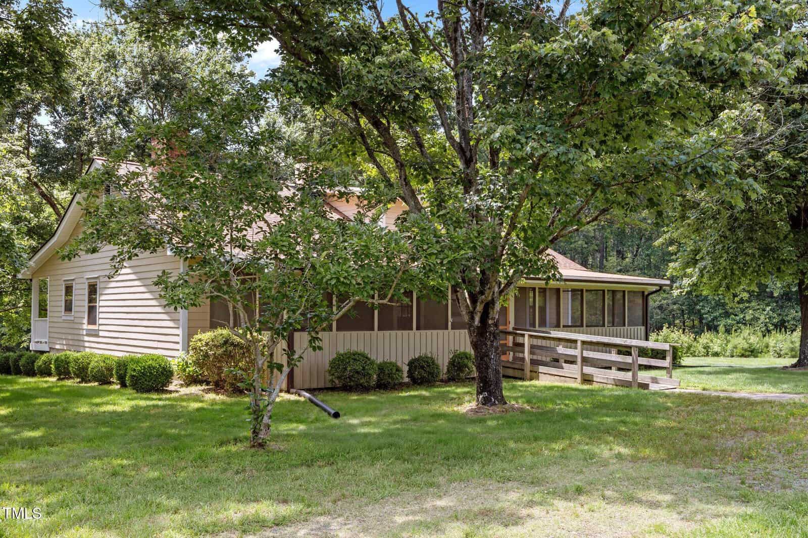 A single-story house with beige siding sits among tall trees. The house has a large screened-in porch, a wooden ramp leading to the entrance, and well-maintained bushes along the side. Grass covers the yard.