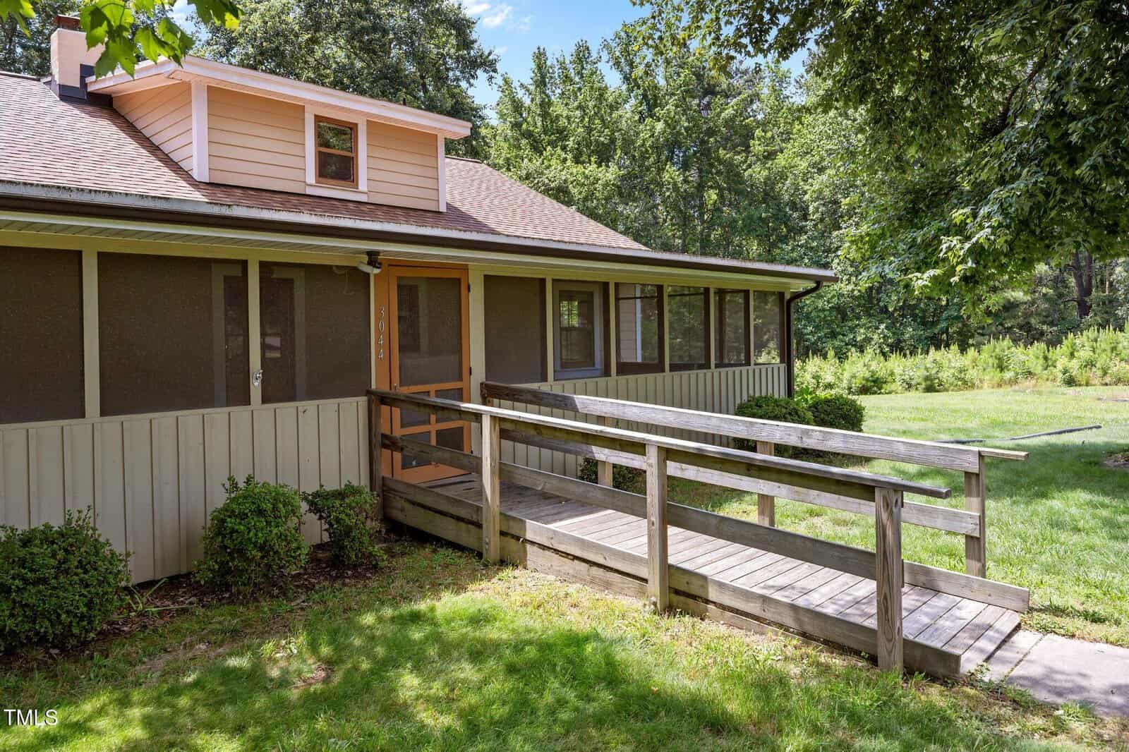 A single-story house with a ramp leading to a screened front porch, surrounded by grass, bushes, and trees under a sunny sky.