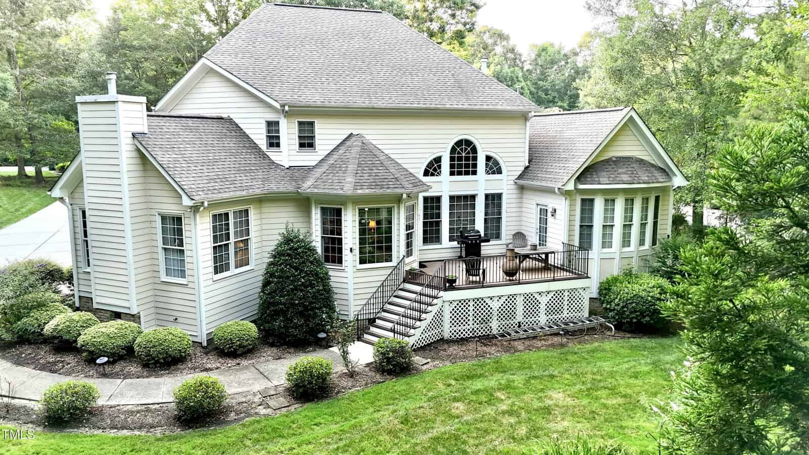 A beige two-story house with multiple peaked roofs, large windows, and a back deck with stairs leading to a landscaped yard bordered by greenery and shrubs.