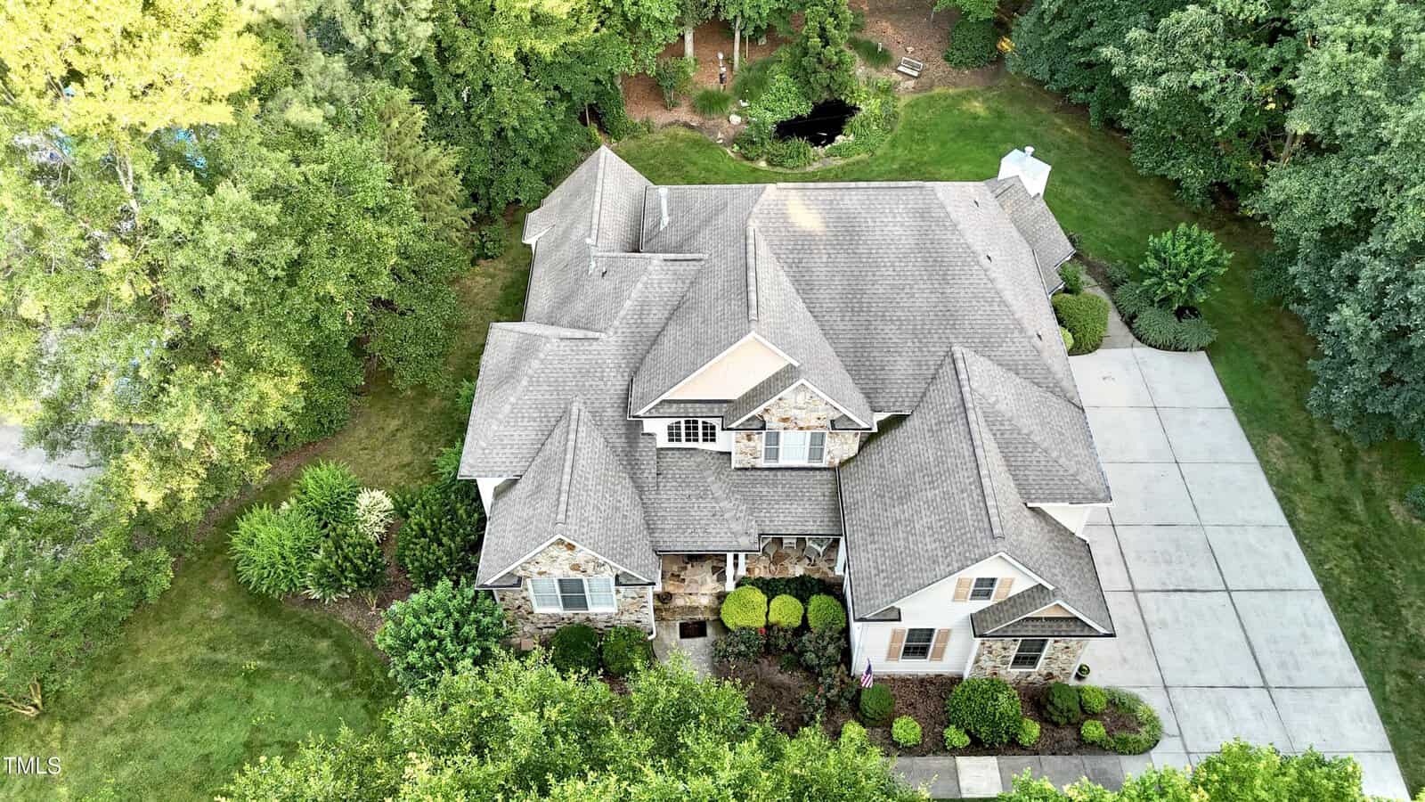 Aerial view of a large two-story house with a stone and siding exterior, gray roof, manicured shrubs, and a spacious driveway, surrounded by green trees and lawn.
