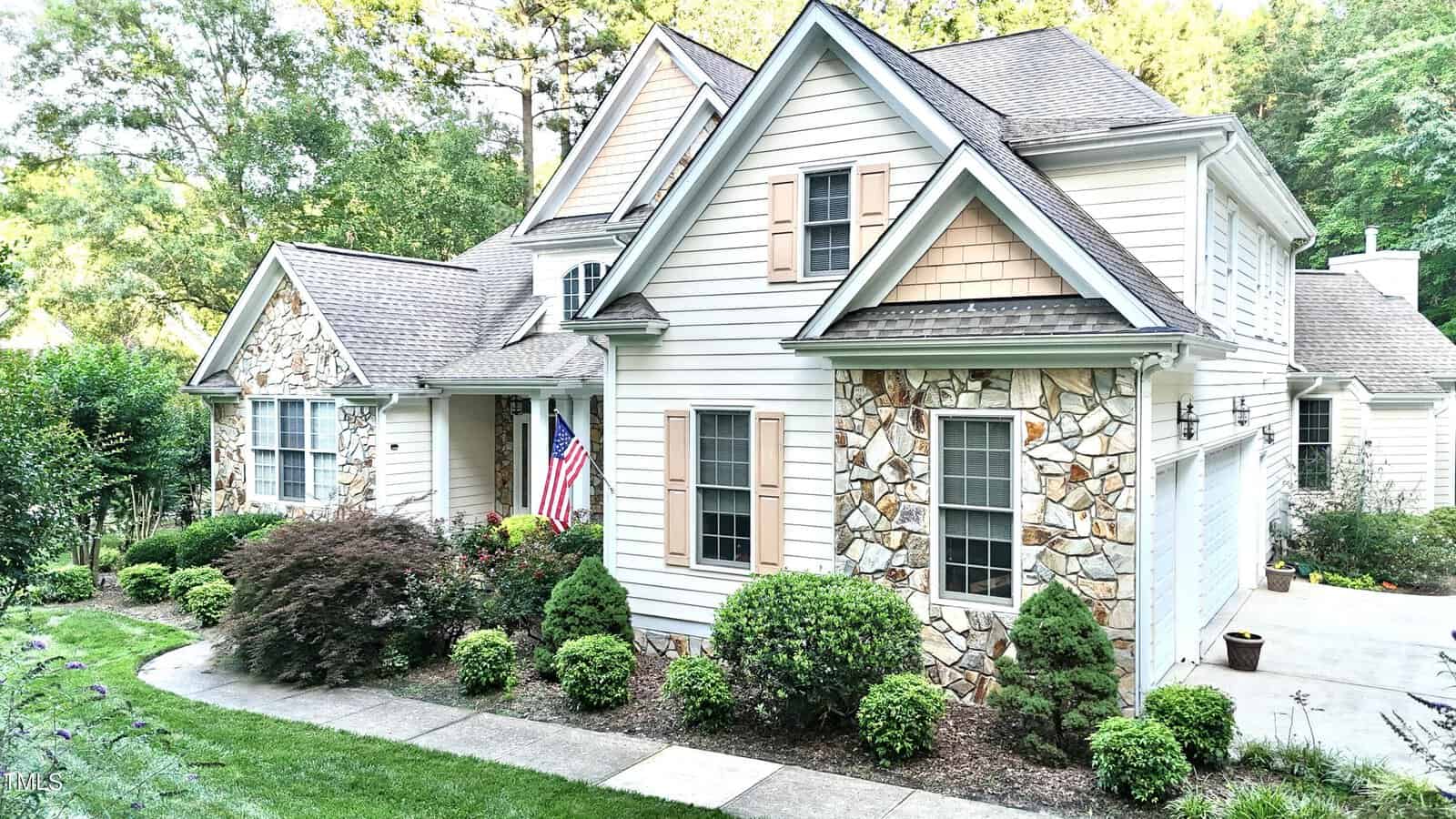 A two-story suburban house with stone and white siding, light brown shutters, a front porch with an American flag, well-kept bushes, and a driveway leading to a two-car garage.