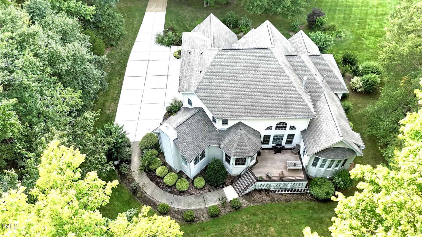 Aerial view of a large two-story house with a gray roof, multiple gables, a wooden deck, landscaped bushes, a curved walkway, and a long driveway surrounded by green lawns and trees.