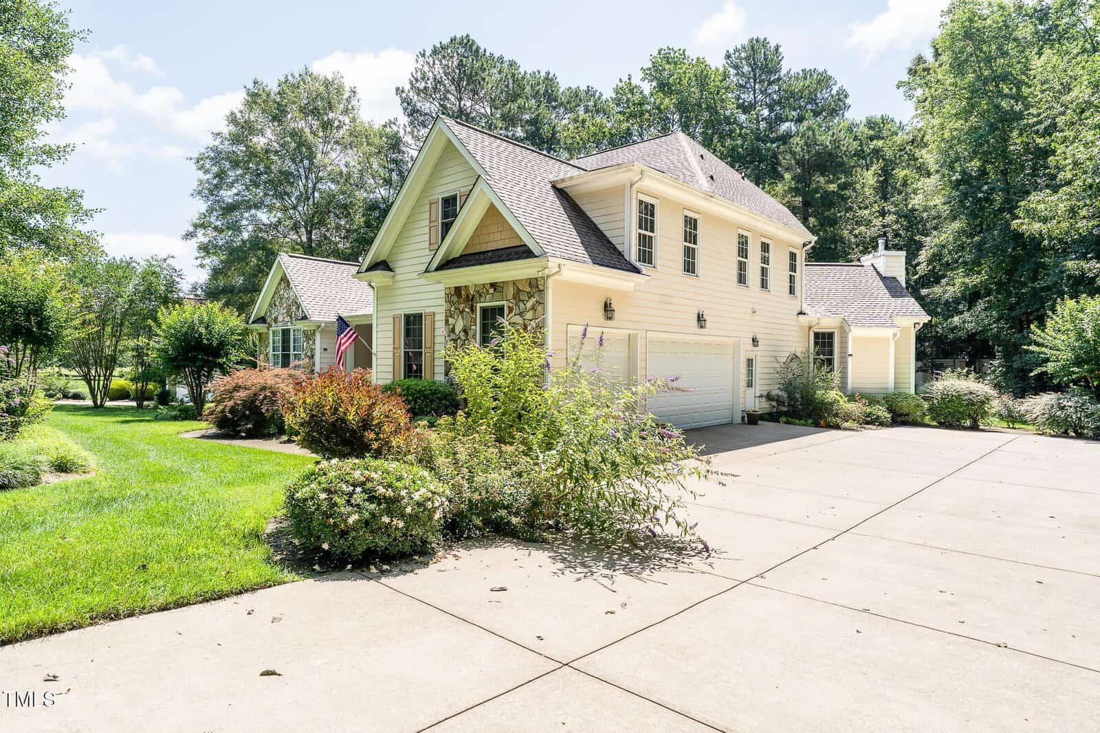 A two-story cream-colored house with a landscaped front yard, an American flag by the porch, attached garage, and a large driveway, surrounded by trees and greenery on a sunny day.
