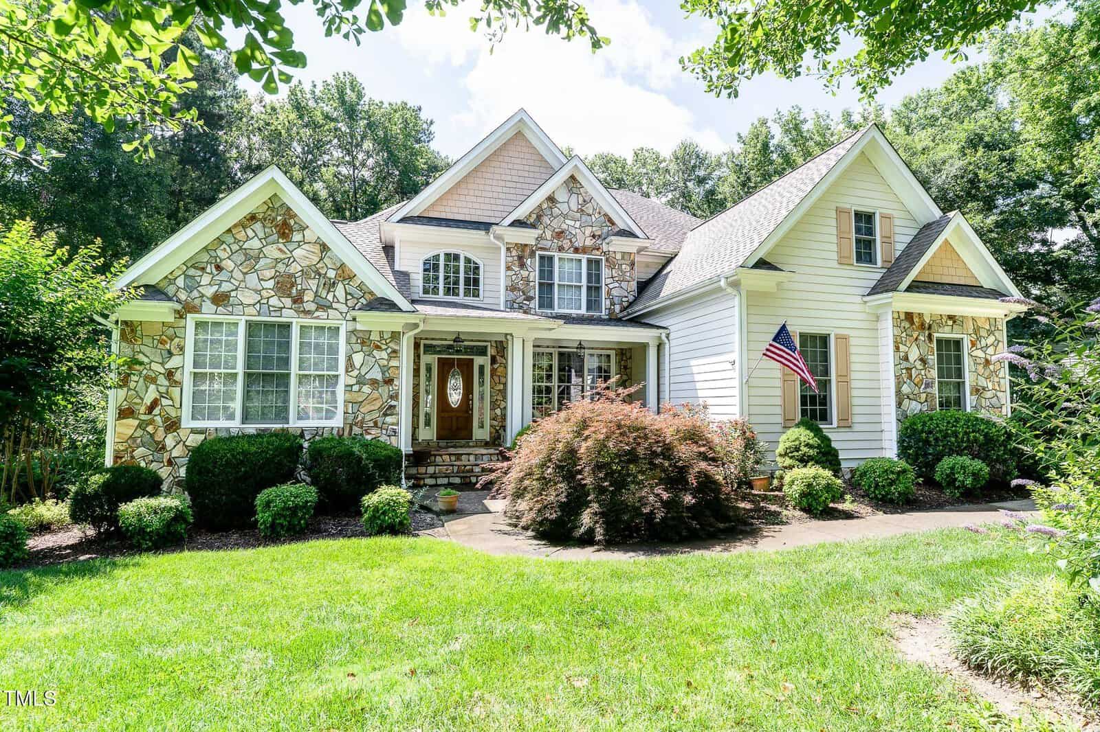 A two-story house with stone and white siding, featuring gabled roofs, a front porch with columns, landscaped bushes, an American flag, and a green lawn under a partly cloudy sky.