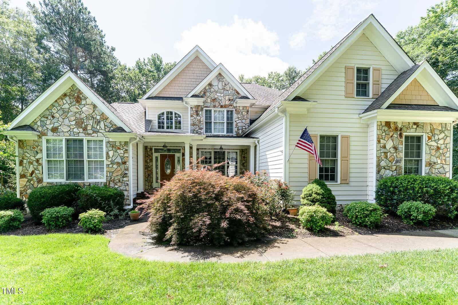 A two-story house with stone and white siding exterior, gabled roof, arched windows, and an American flag displayed near the entrance. Lush green lawn, shrubs, and trees surround the front yard.