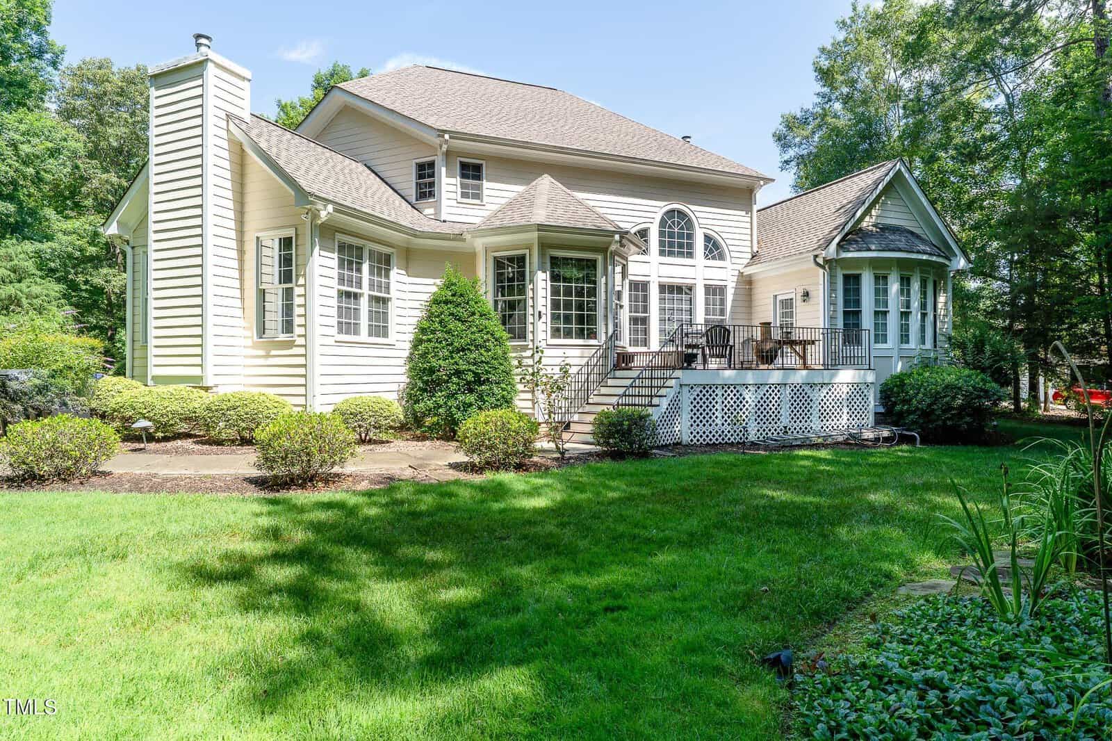 A two-story house with beige siding, large windows, a spacious back deck with railings and stairs, surrounded by green lawn, shrubs, and trees on a sunny day.