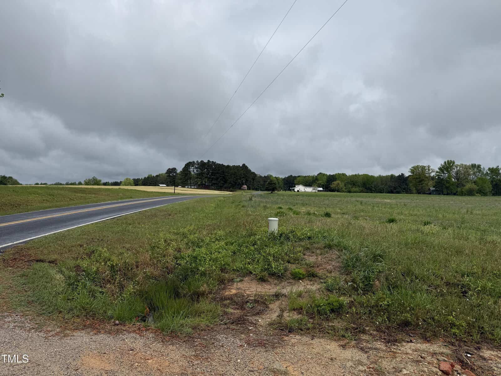 A rural scene featuring a paved road curving left, open grassy fields on both sides, scattered trees in the background, and a cloudy sky overhead. A small white utility box is visible near the roadside.