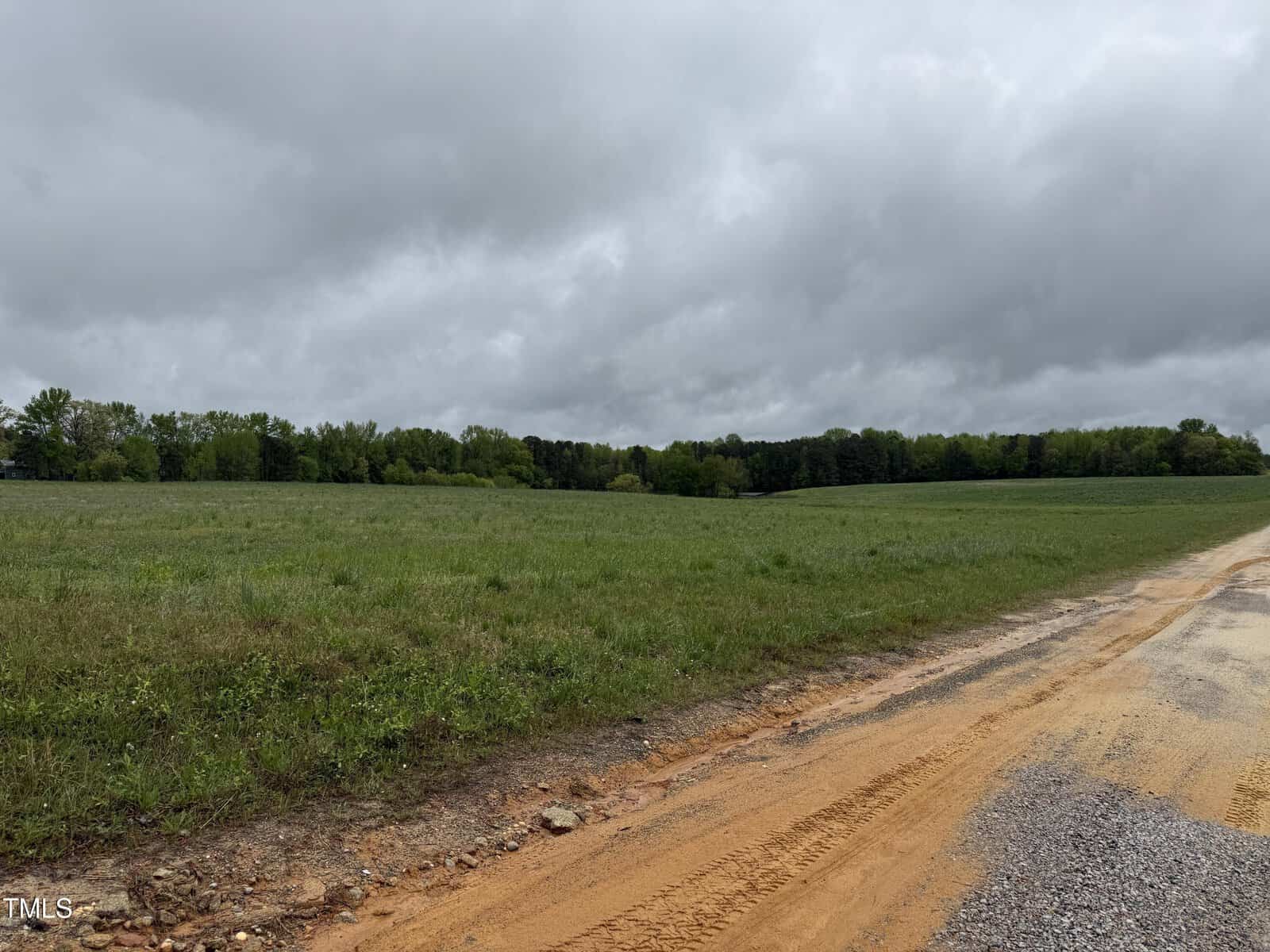 A cloudy sky hangs over a wide, grassy field bordered by trees, with a reddish dirt and gravel road running along the right side of the image.