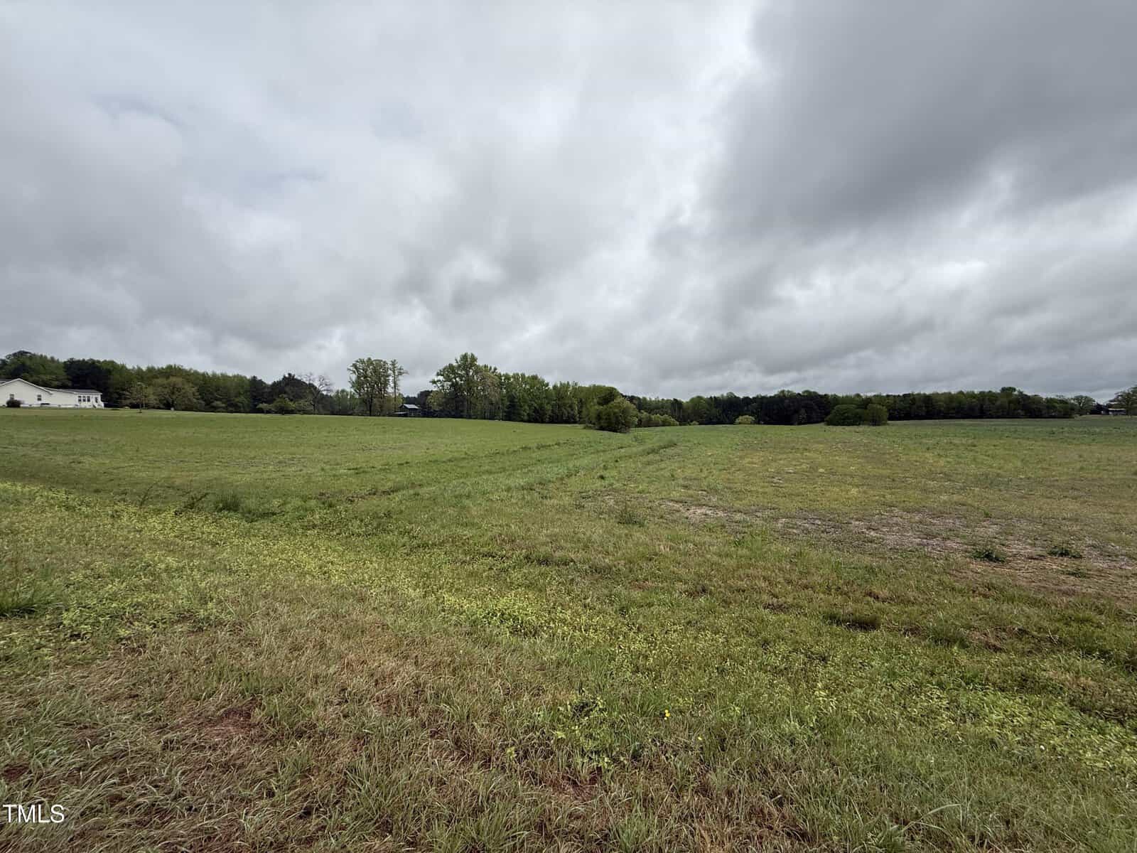 A large open grassy field under a cloudy sky, bordered by trees in the distance. A house is partially visible on the far left side. The landscape appears flat and rural.