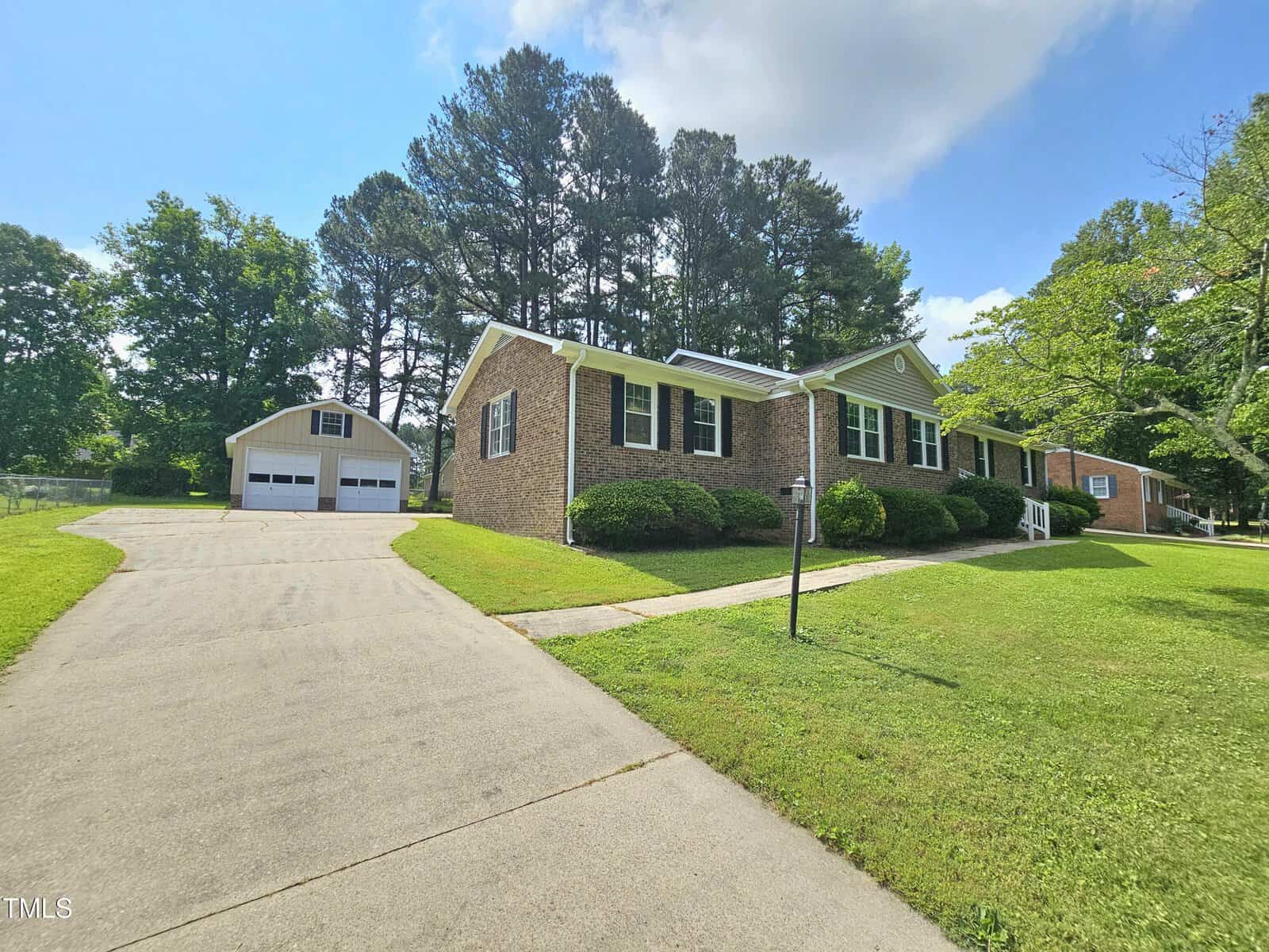 Single-story brick house with white trim, a well-kept lawn, and a wide driveway leading to a detached two-car garage, surrounded by green trees under a partly cloudy sky.