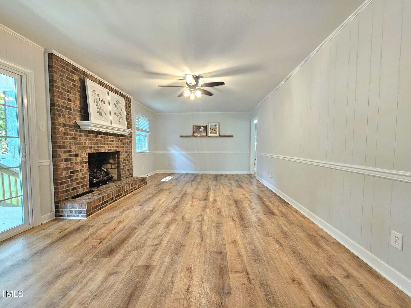 Spacious living room with wood flooring, brick fireplace, white paneled walls, ceiling fan with lights, two wall shelves, and large windows letting in natural light.