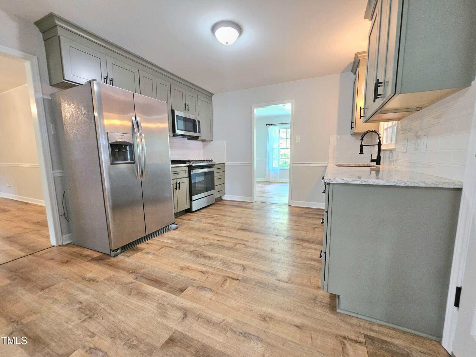 Modern kitchen with light wood flooring, gray cabinets, stainless steel appliances, and white backsplash. An open doorway leads to a bright room with large windows in the background.