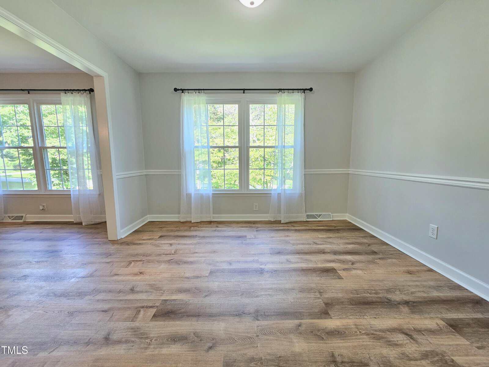 Bright, empty room with large windows, sheer white curtains, light gray walls, and wood flooring. Sunlight streams in, and an adjoining room with matching decor is visible through an open doorway.