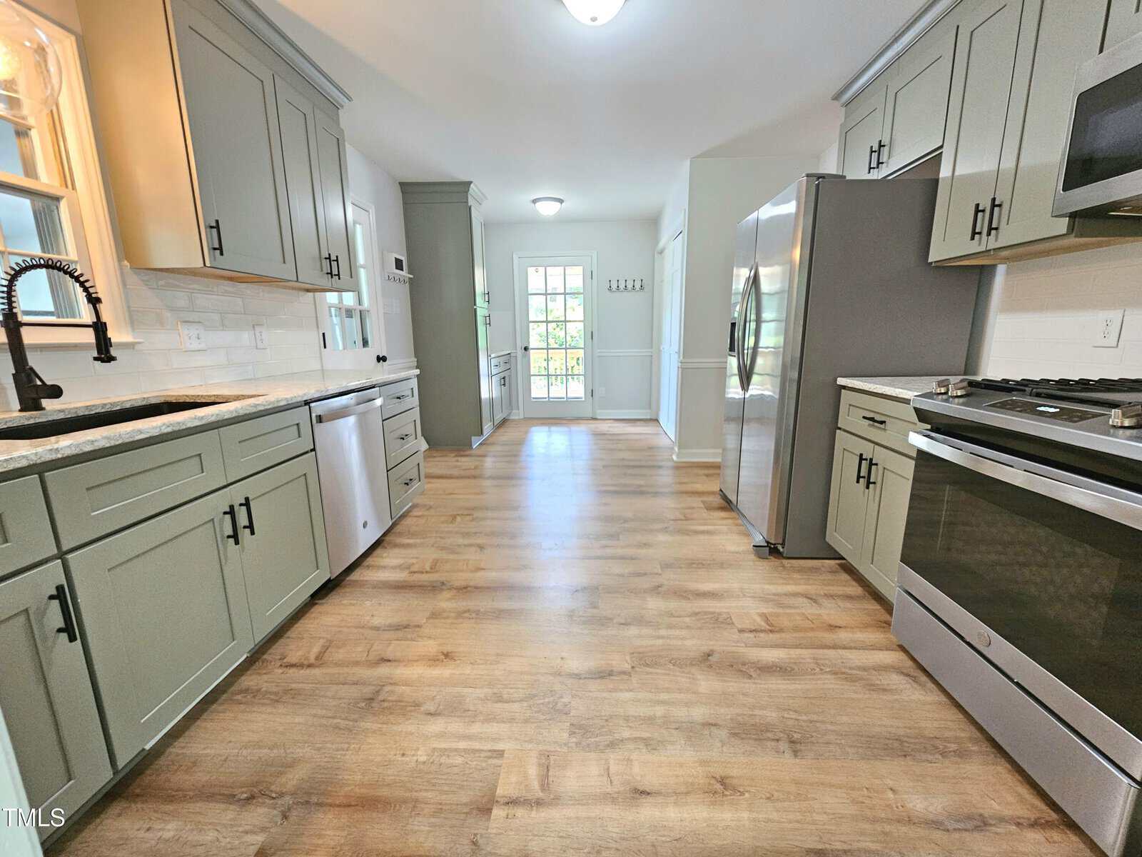A modern kitchen with green cabinets, stainless steel appliances, light countertops, wood-style flooring, and a view of double doors leading to a bright, sunlit room at the far end.