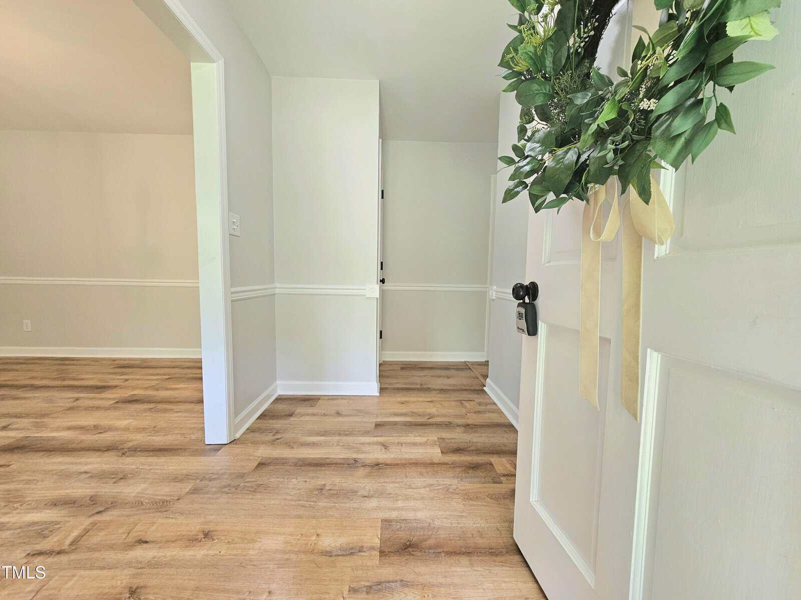 A white door with a green wreath and beige ribbon is open to a room with light-colored walls, white trim, and wood-style flooring. The room is empty and well-lit with natural light.