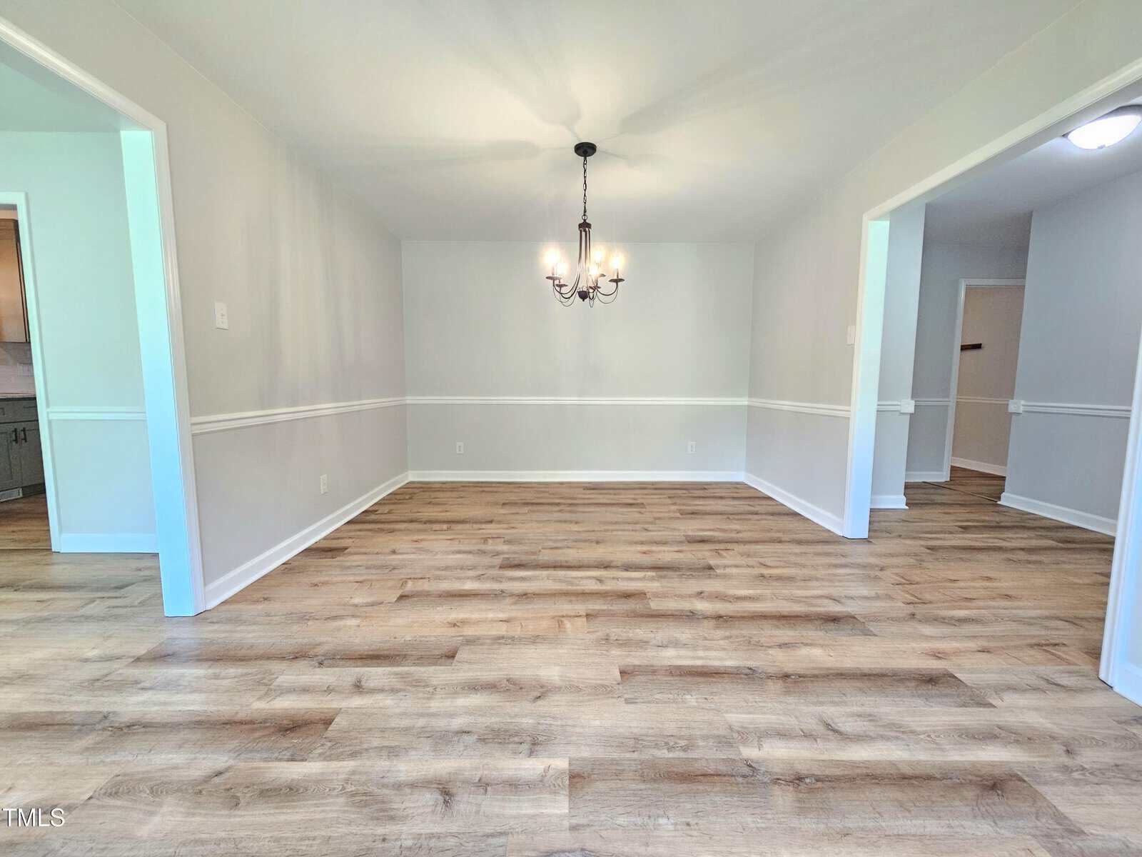 A bright, empty dining room with light-colored walls, wood flooring, white trim, and a black chandelier. Open doorways lead to other rooms, one with visible kitchen cabinets and another with a hallway light.