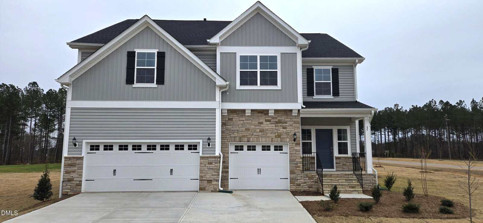 Located in Youngsville at 85 Guardian Street, this two-story house features gray siding, stone accents, three garage doors, and a covered front porch. The lawn is brown, with small shrubs near the entrance. Trees and an open field are visible in the background.