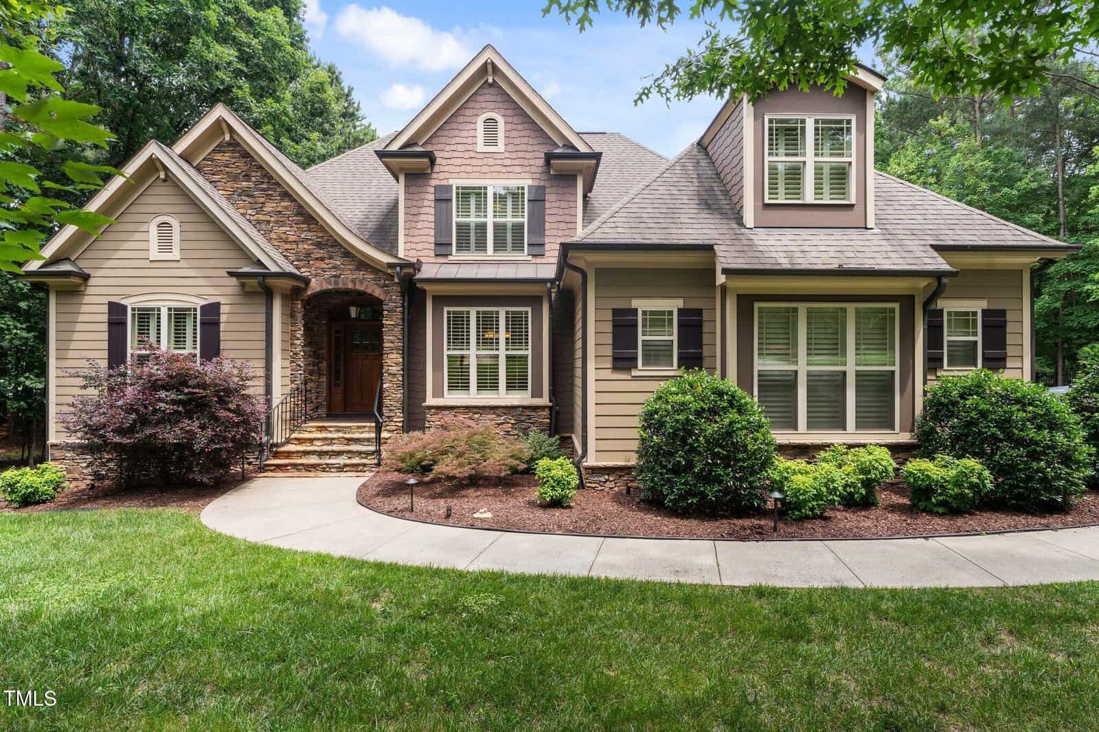 A tan two-story house with stone accents at 512 Belmont Circle, Wake Forest, features black shutters, white window trim, and sits behind a curved sidewalk surrounded by manicured shrubs, small trees, and green grass with tall trees in the background.